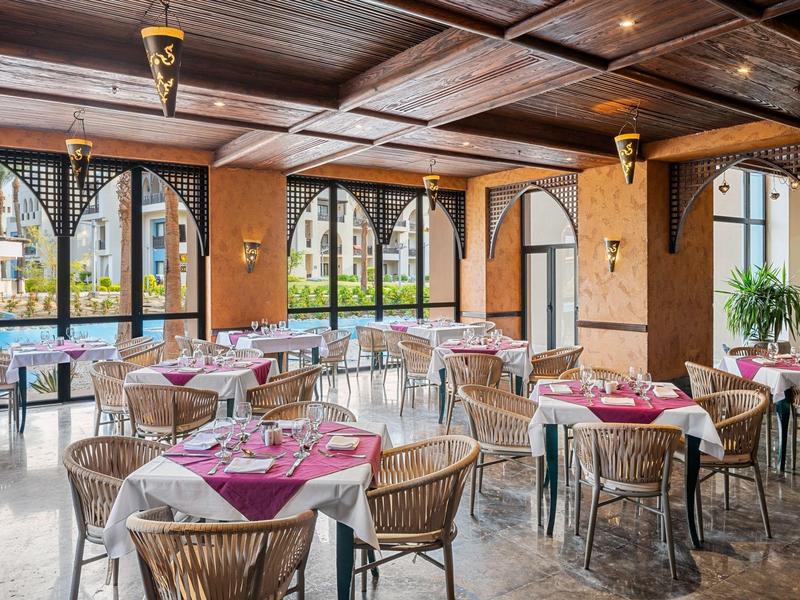 Světla jídelna s kulatými stoly, červenými ubrusy a velkými okny.