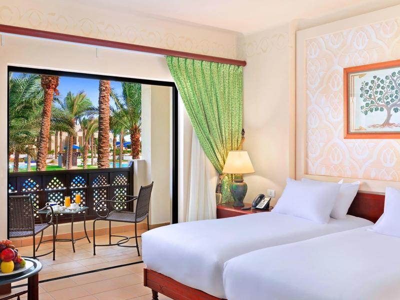 Světle zařízený hotelový pokoj se dvěma postelemi, balkonem a výhledem na palmy.