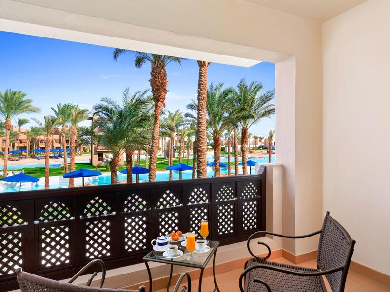 Hotelová terasa se stolem a židlemi, výhled na bazén a palmy pod modrou oblohou.