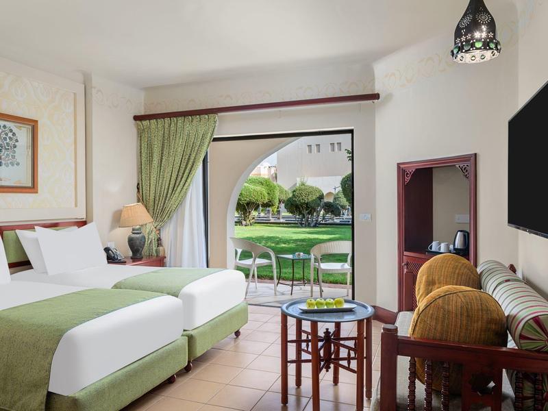 Světelný hotelový pokoj se zelenými dvojlůžky a terasou s výhledem do zahrady.