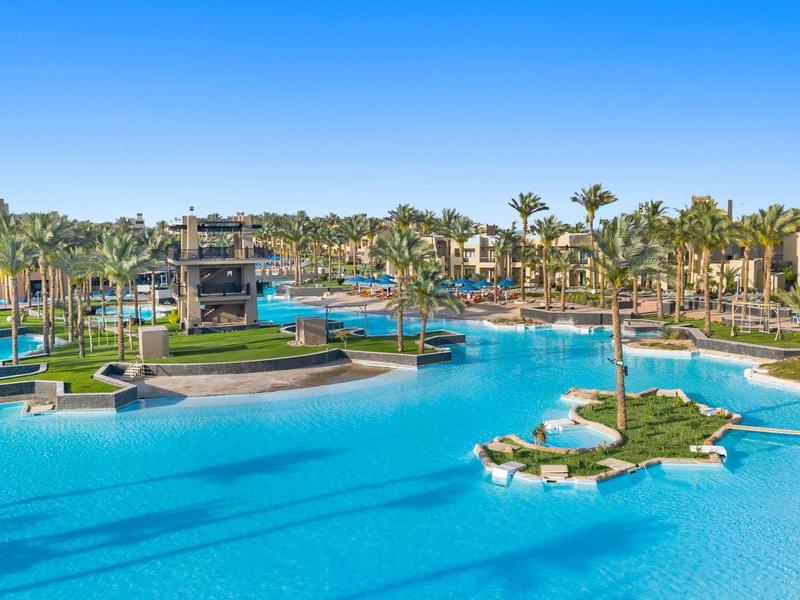 Hotelový bazén s ostrůvky a palmami pod jasnou modrou oblohou.