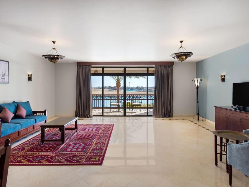 Lichte woonkamer met groot raam uitzicht op water, blauwe en rode meubels en tapijt.