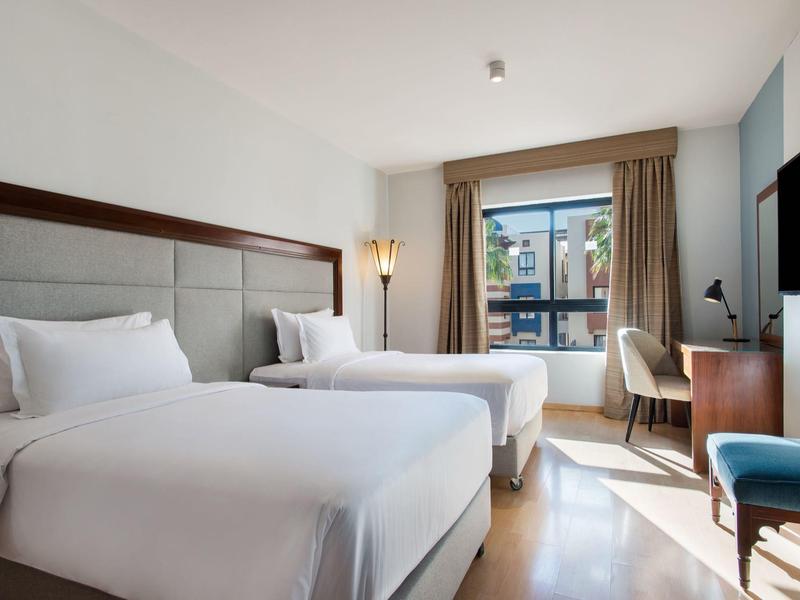 Lichte hotelkamer met twee eenpersoonsbedden, bureau en balkon met uitzicht naar buiten.