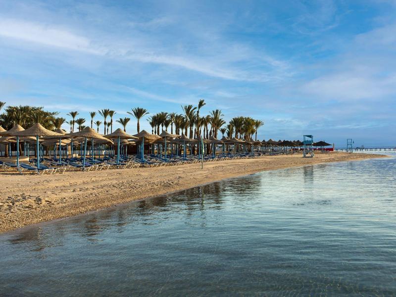 Een rustige zandstrand met palmbomen, parasols en hutten onder een blauwe lucht.