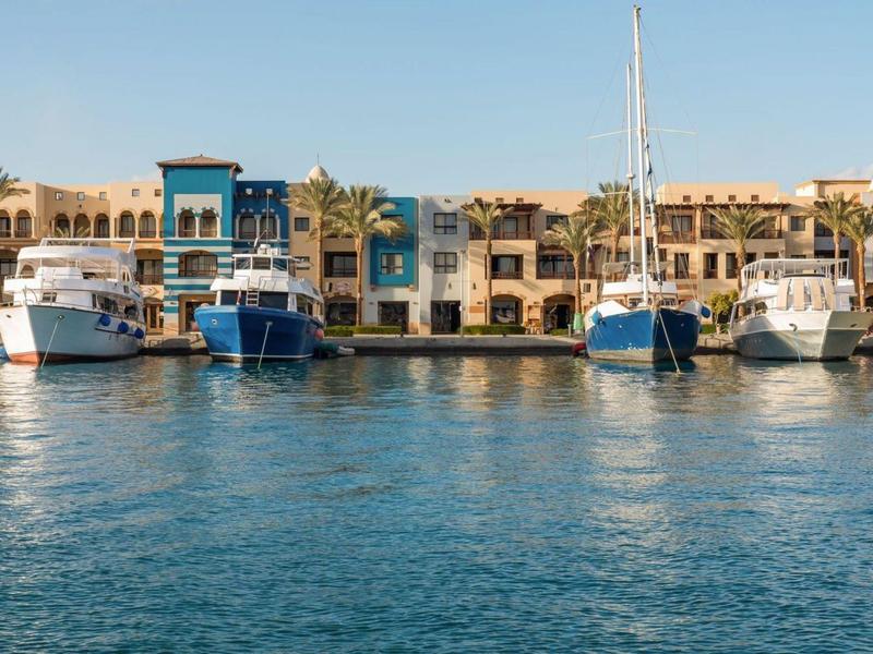 Jachten aangemeerd aan de kade voor kleurrijke gebouwen en palmbomen onder een heldere lucht.