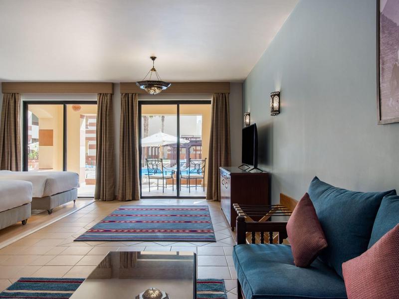 Lichte hotelkamer met bed, bank, tapijten en terras met uitzicht op het zwembad.