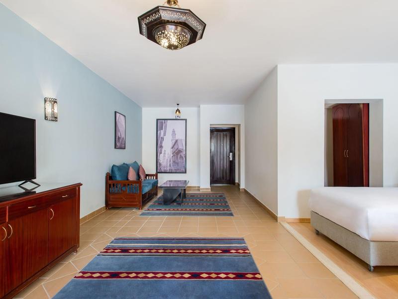 Lichte hotelkamer met groot bed, tv op houten kast, zithoek en kleurrijk tapijt.