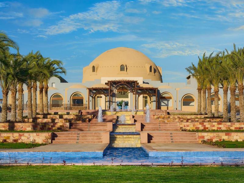 Große weiße Gebäude mit Kuppel, umgeben von Palmen, Treppen und blauem Wasserbecken bei klarem Himmel.
