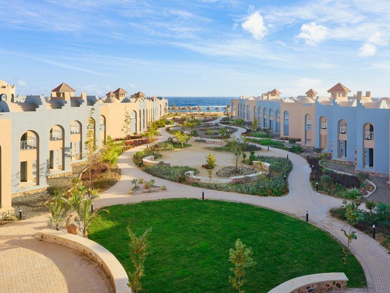 Blick auf weiße Häuser mit roten Dächern und Wegen, umgeben von grünem Garten unter blauem Himmel.