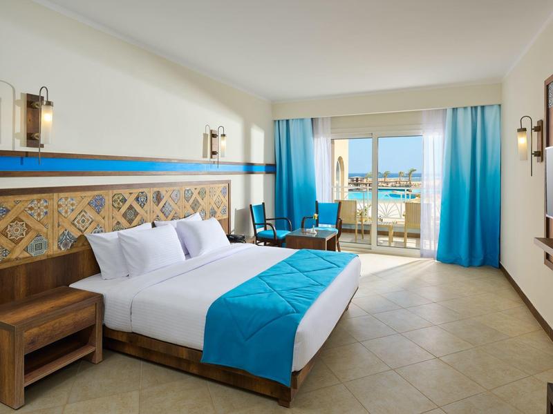 Helle Hotelzimmer mit Doppelbett, blauen Akzenten, Balkon und Meerblick.