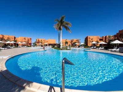 Runder Hotelpool mit blauem Wasser und Palmen, umgeben von Liegestühlen bei sonnigem Himmel.