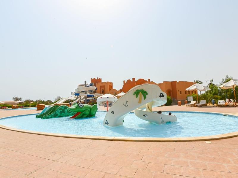 Plac zabaw dla dzieci z zjeżdżalnią wodną i sprzętem w płytkim basenie przed budynkami hotelu.