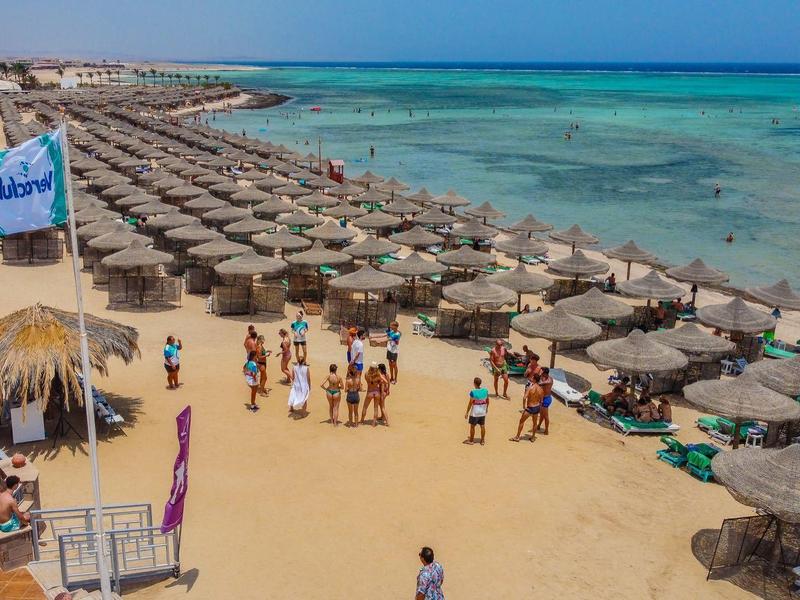 Plaża z rzędami słomianych parasoli, ludźmi na piasku i czystym niebieskim morzem w tle.
