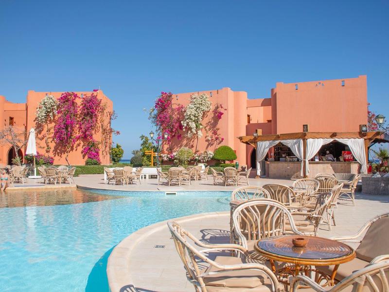 Widok na hotelowy basen z okrągłymi stołami i krzesłami pod jasnym niebieskim niebem.