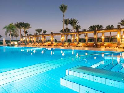 Piscina esterna illuminata con palme e lettini in un hotel al crepuscolo.