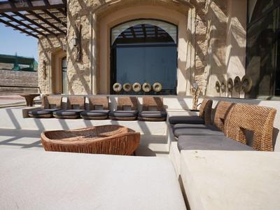 Buitenzitgedeelte met houten tafels en beklede stoelen naast zandgrond.