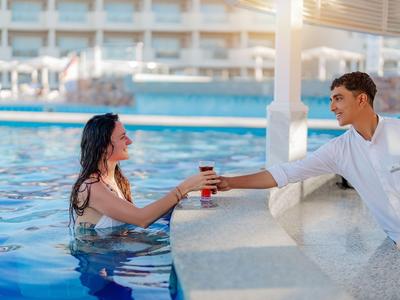 Donna in piscina che riceve una bevanda da un uomo con camicia bianca al bar della piscina.