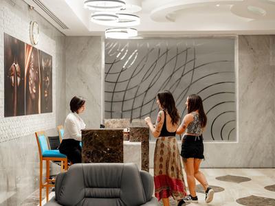 Réception d'hôtel avec un comptoir en marbre, deux clientes parlant à la réceptionniste, et des œuvres d'art aux murs.