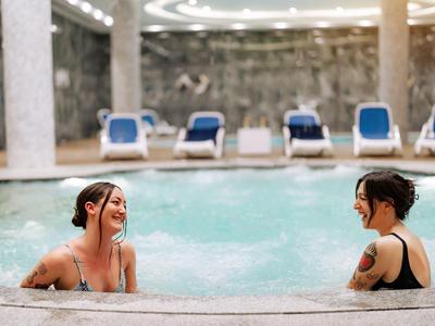 Deux femmes profitent de la baignade dans une piscine intérieure avec des chaises longues en arrière-plan.