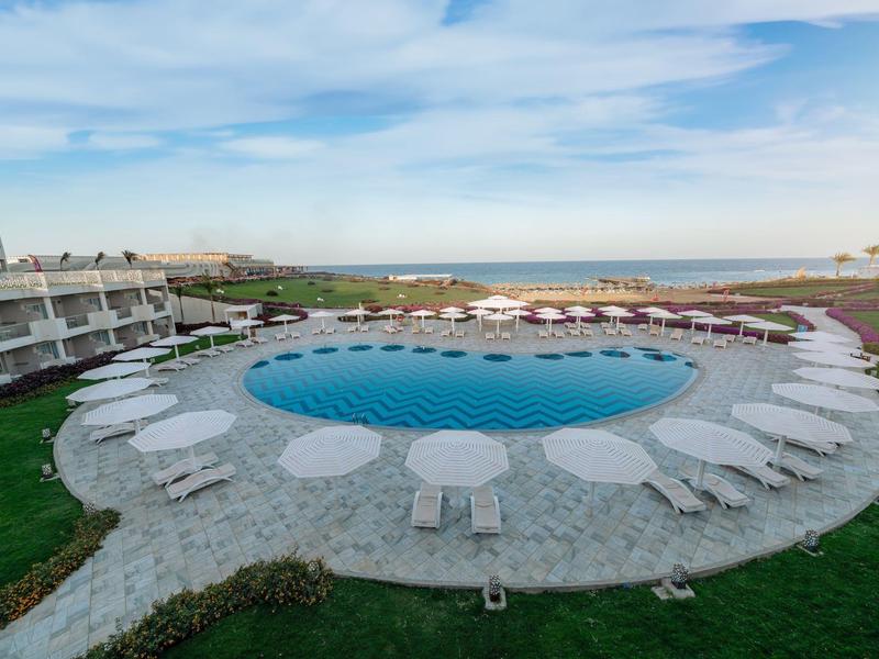 Piscine ronde entourée de transats et parasols sous un ciel bleu près d'un hôtel.