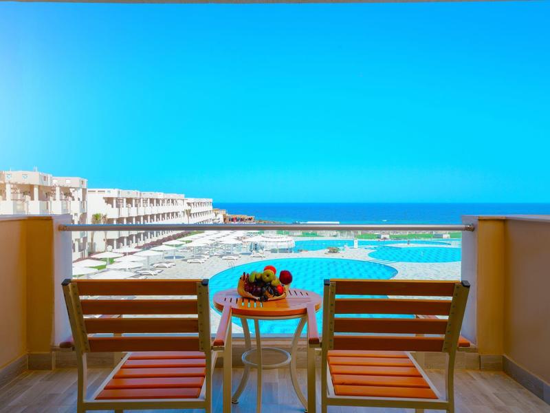 Balcon avec deux chaises et une table donnant sur la piscine du complexe et l'océan sous un ciel bleu clair.