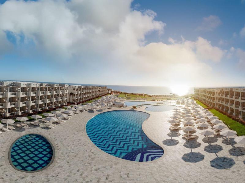 Coucher de soleil sur une station balnéaire avec bâtiments, parasols et piscine entre les zones sablonneuses.