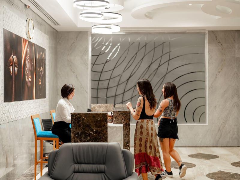 Réception d'hôtel avec un comptoir en marbre, deux clientes parlant à la réceptionniste, et des œuvres d'art aux murs.