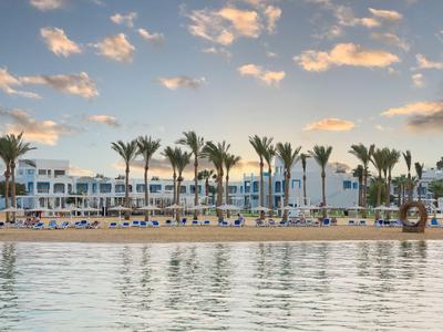 Hotel na pláži s palmami a klidnou vodou při západu slunce.