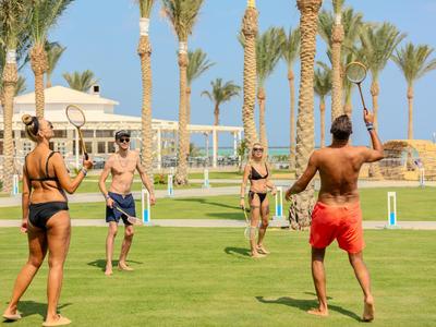 Skupina lidí hrající volejbal na travnaté ploše s palmami v pozadí.