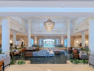 Prostorná hotelová lobby s bílým interiérem, sloupy, lustr a výhledem na moře v pozadí.