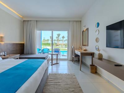 Moderní hotelový pokoj s manželskou postelí, psacím stolem, TV a výhledem na terasu a palmy.