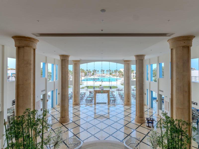 Světlá hotelová hala se sloupy, velkým oknem a výhledem na bazén.