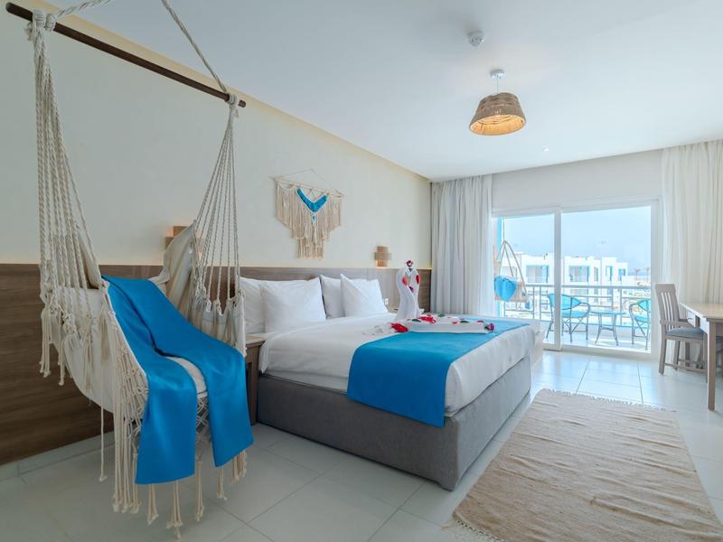 Moderní hotelový pokoj s manželskou postelí, závěsným křeslem a balkonem s výhledem na moře.