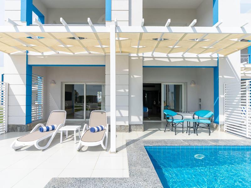 Moderní terasa s bazénem, lehátky a jídelním stolem před bílou budovou.