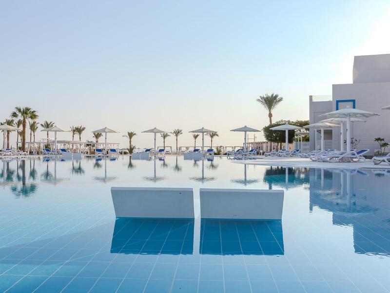 Infinity bazén s lehátky a palmami na hotelové pláži pod jasnou oblohou.