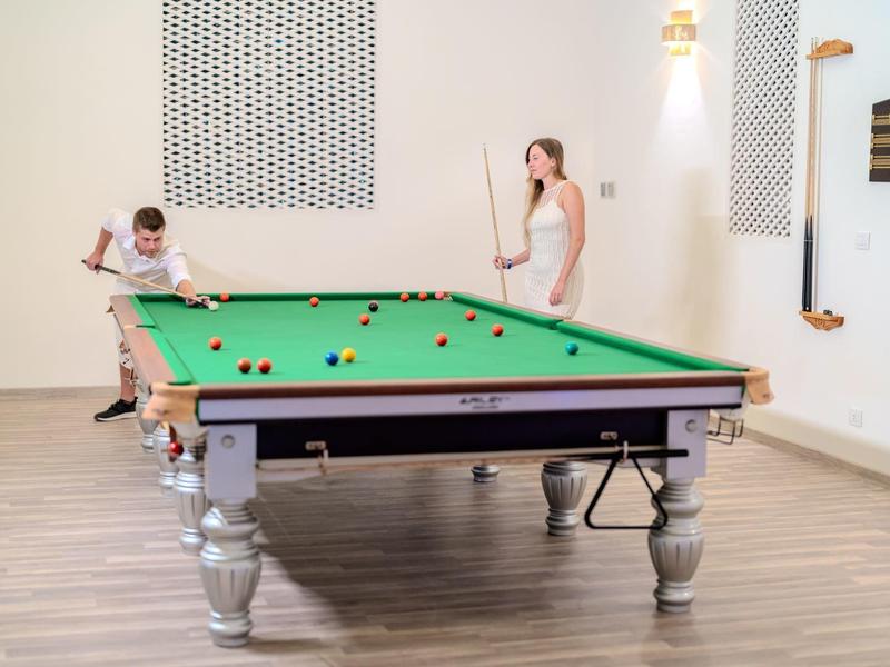 Dvě ženy hrají kulečník v světlé místnosti s dřevěnou podlahou a bílými stěnami.