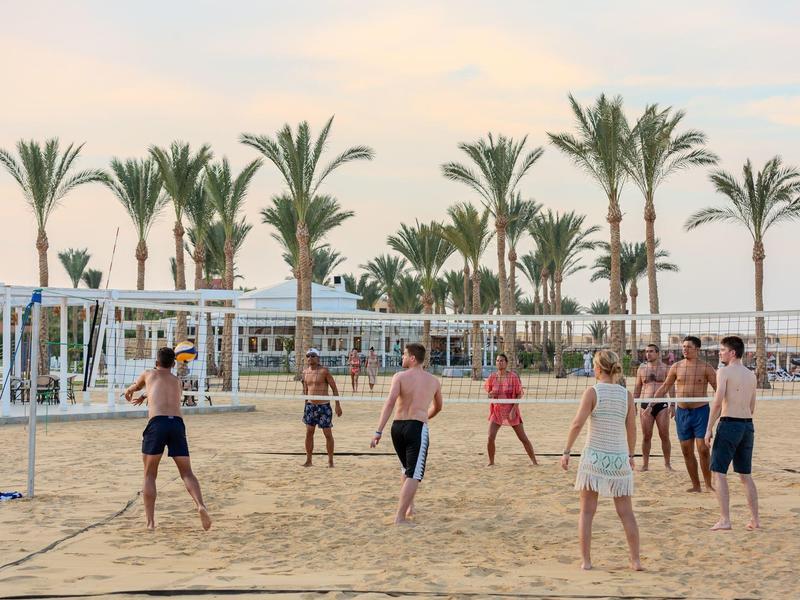 Lidé hrají volejbal na pláži s palmami v pozadí při západu slunce.