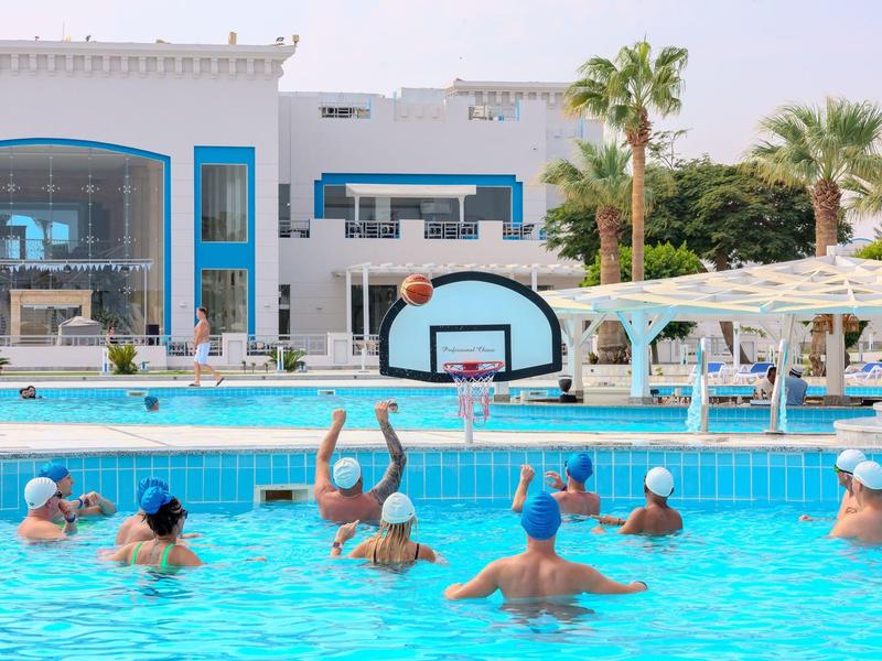 Skupina lidí hraje vodní basketbal v hotelovém bazénu s moderní fasádou.