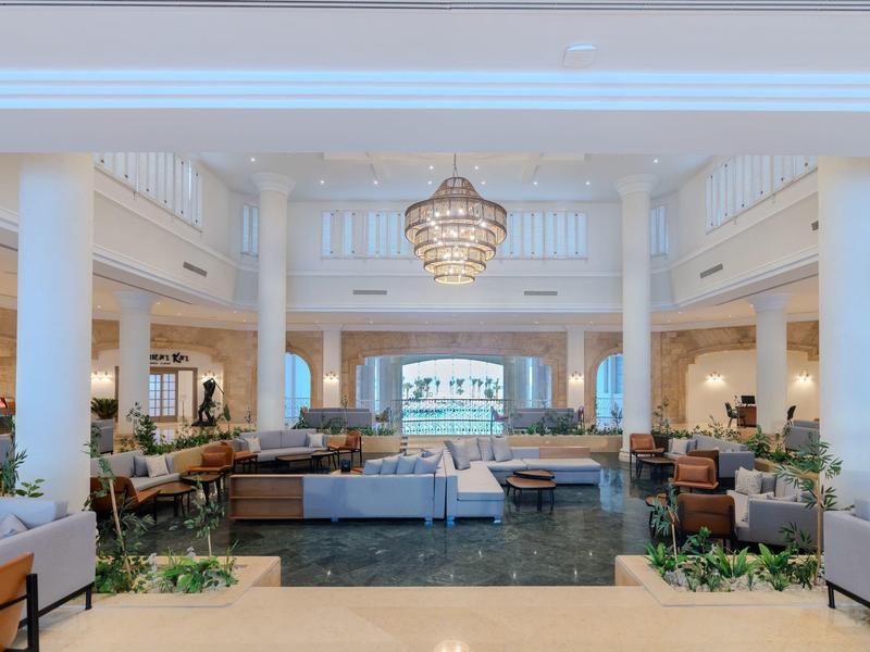 Prostorná hotelová lobby s bílým interiérem, sloupy, lustr a výhledem na moře v pozadí.
