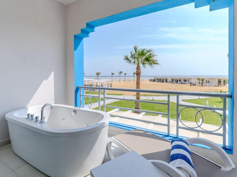 Volně stojící vana na balkoně s výhledem na moře a palmy v pozadí.