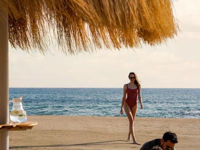 Strandszene mit Liegestuhl, Strohdach und Frau im roten Badeanzug.