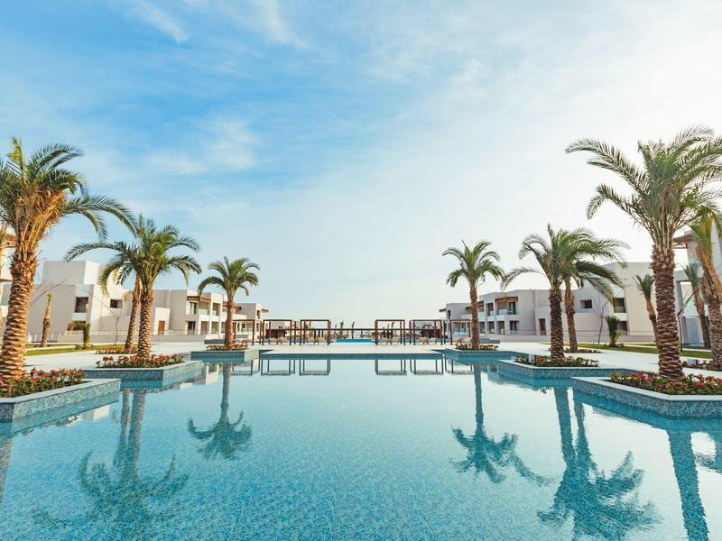 Piscina azzurra limpida con palme e edifici bianchi sotto un cielo luminoso.