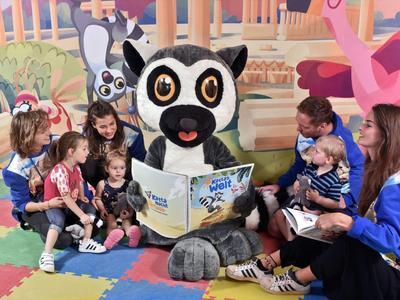 Kinder sitzen auf buntem Spielteppich, schauen mit zwei Betreuern und Lemuren-Maskottchen ein Buch an.