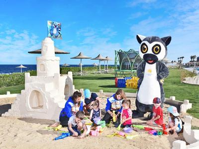 Kinder spielen am Strand in einem Sandkasten, neben einer großen Sandburg mit Meer und blauem Himmel im Hintergrund.