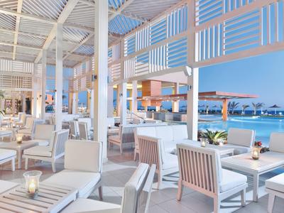 Offene Strandbar mit weißen Möbeln, Kerzen und Meerblick unter hellem Himmel.