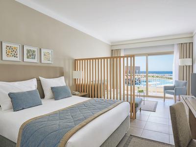 Modernes Hotelzimmer mit Doppelbett, blauen Kissen, Holzteiler und Meerblick durch große Fenster.