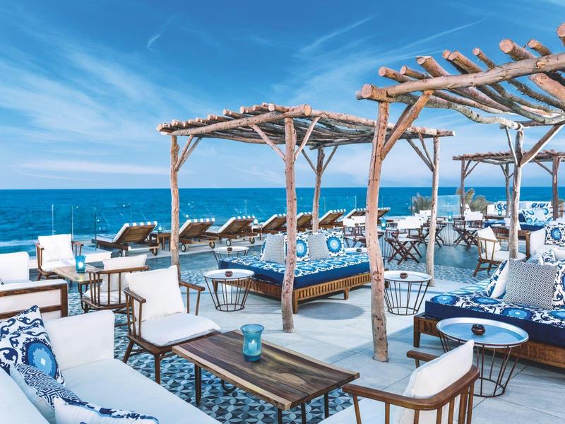 Terraza frente al mar con sofás y mesas bajo pérgolas de madera para descanso junto al océano.