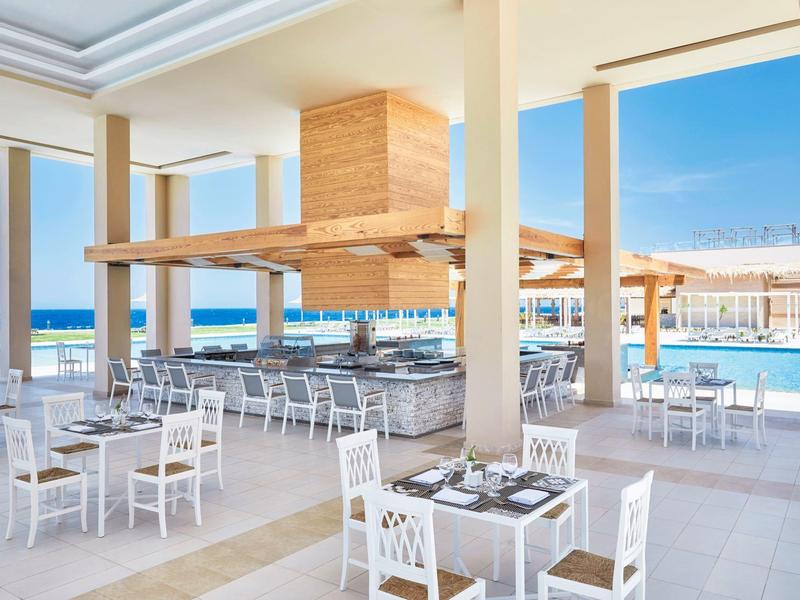 Restaurante abierto con mesas y sillas blancas junto a una piscina con vista al mar.