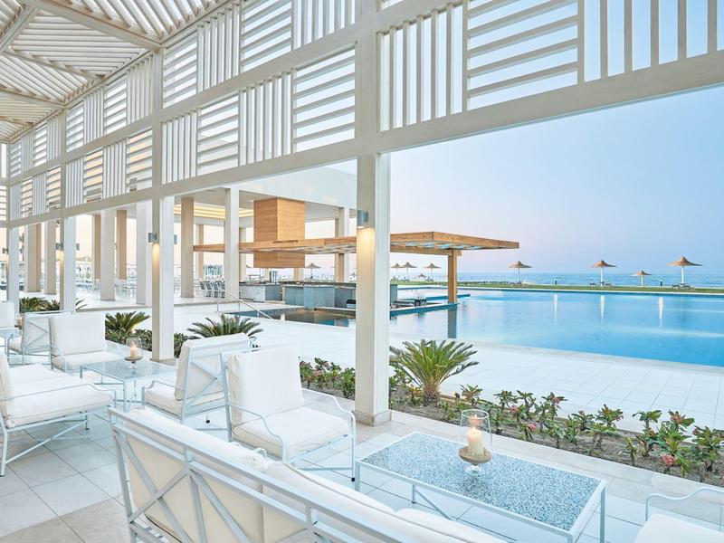 Área moderna de piscina exterior con mobiliario blanco bajo pérgola y vista al mar con cielo despejado.