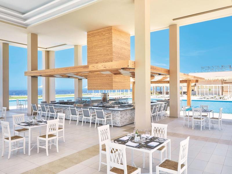 Área de comedor al aire libre con mesas, sillas blancas y vista a la piscina y al mar.